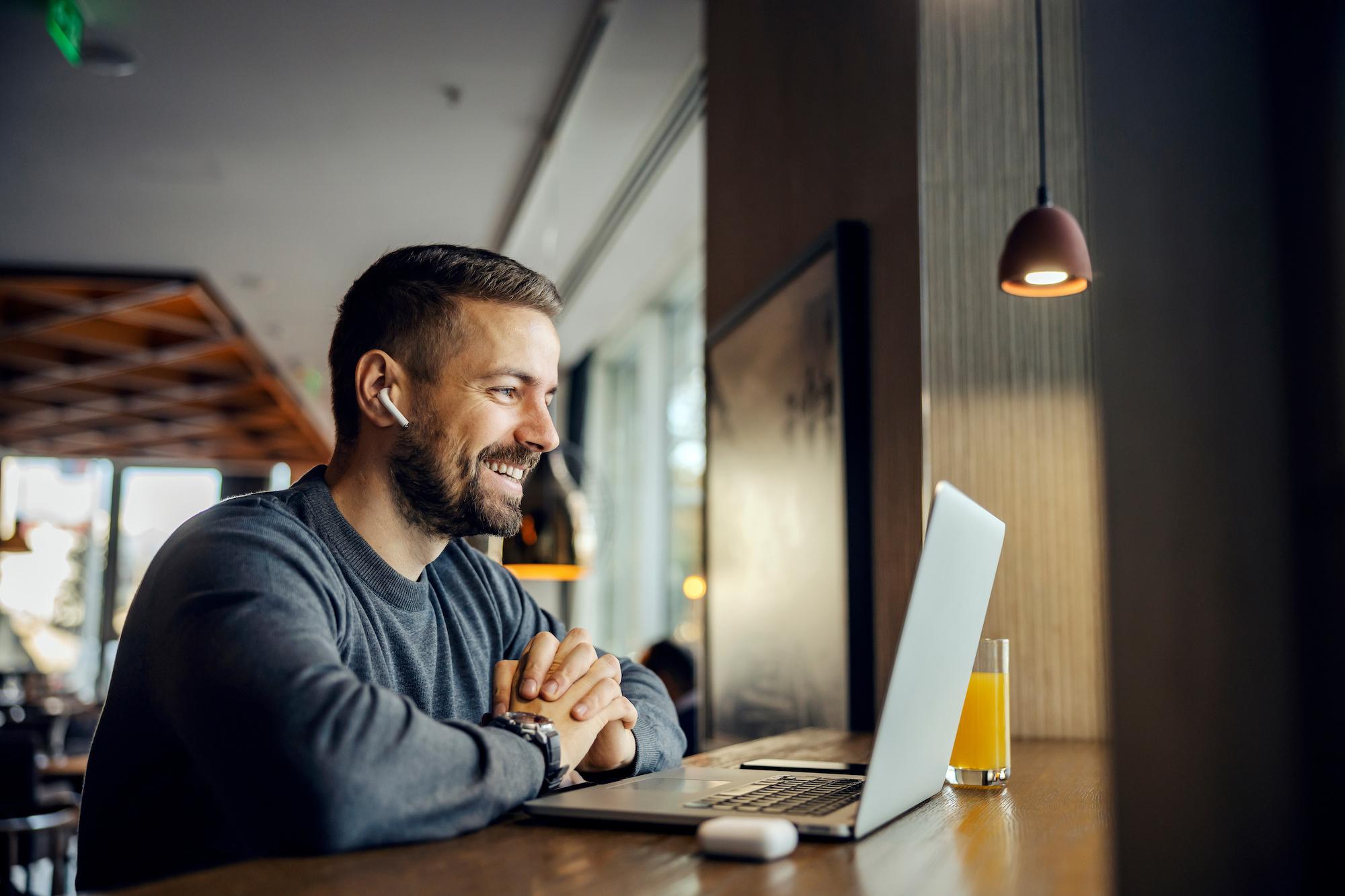 A happy remote worker is sitting in cafeteria and having an online meeting with his clients.